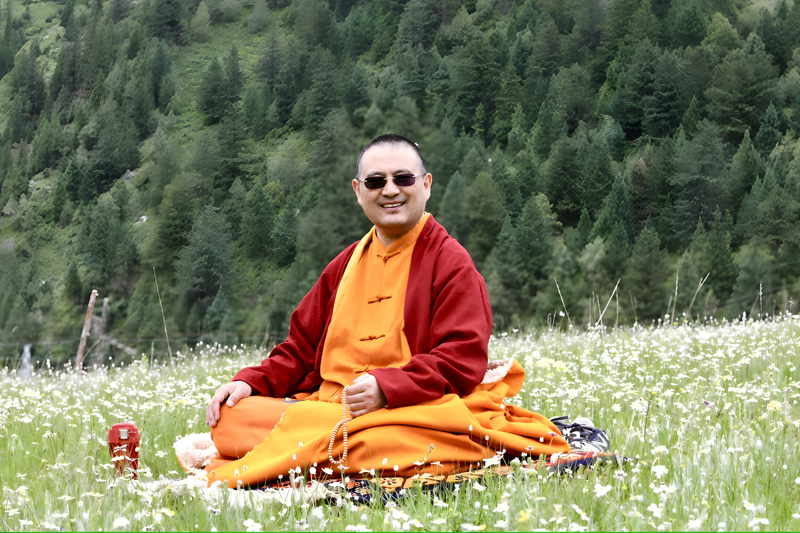 A Buddhist teacher in an orange-red robe seated in a meadow of white flowers