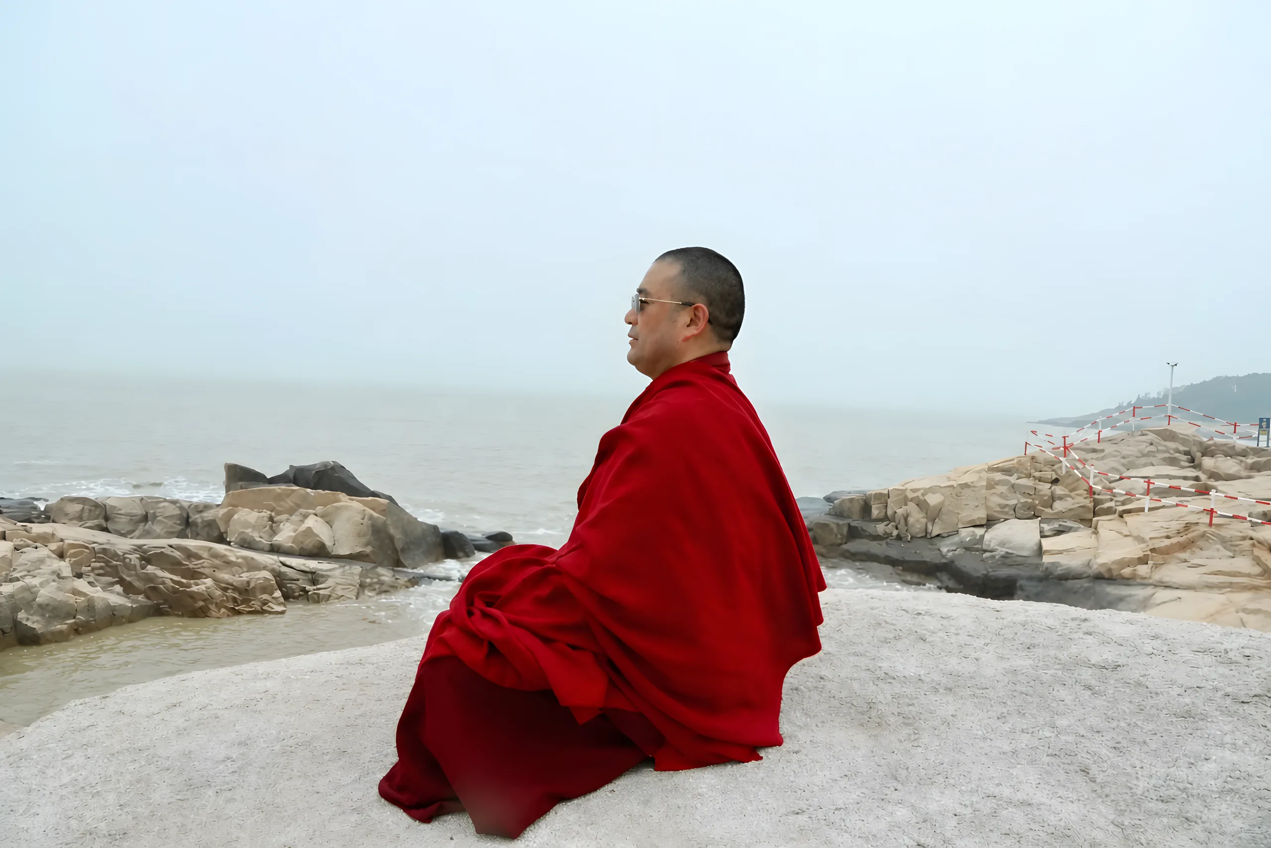 A Buddhist teacher in a red robe seated beside the sea